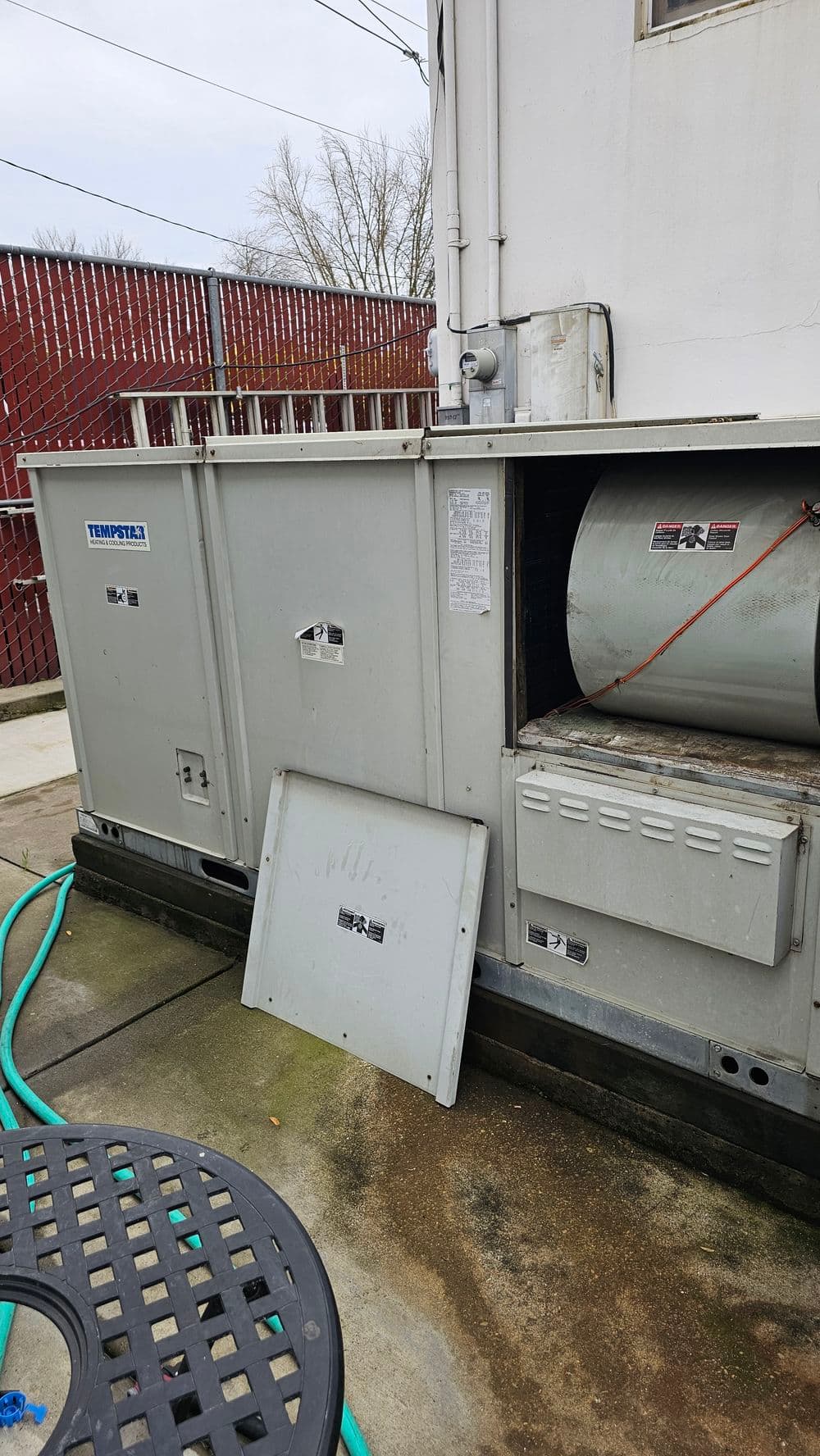Commercial HVAC unit with a disassembled panel and surrounding garden hose on a patio.