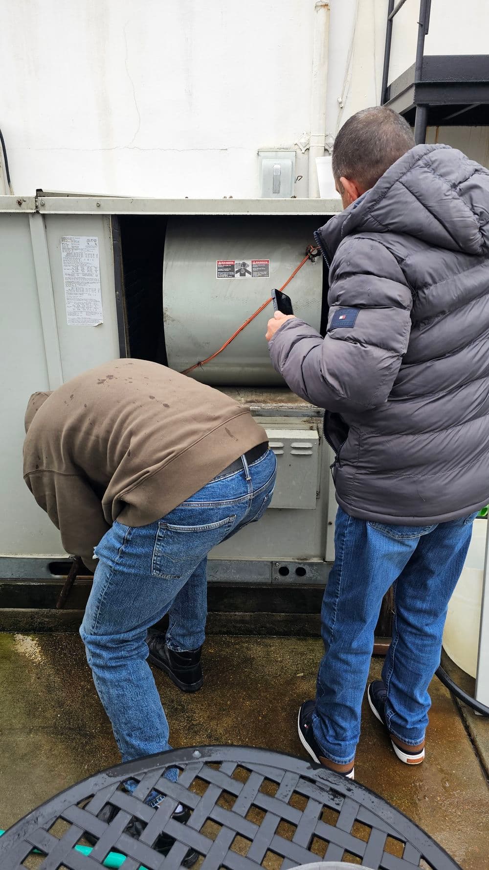 Two men inspecting an outdoor HVAC unit, one using a phone for assistance.